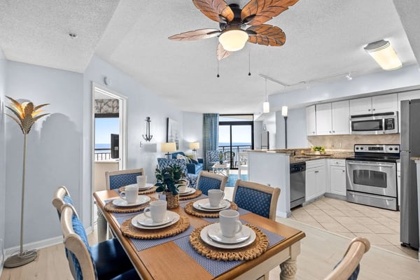 Open-plan coastal condo with dining table set for four, kitchen with stainless steel appliances, and ocean view through glass doors.