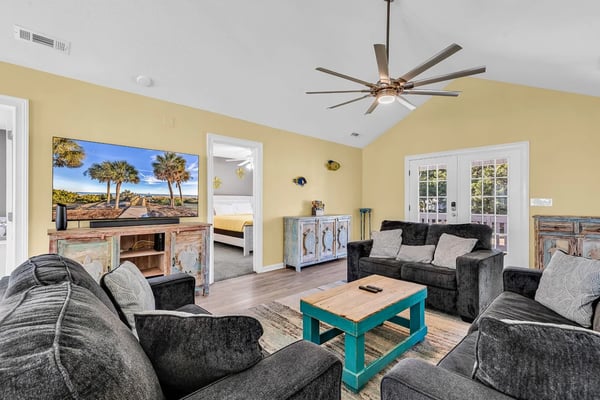 Beachfront condo living room with gray seating, ocean view balcony, and bedroom visible through open doorway